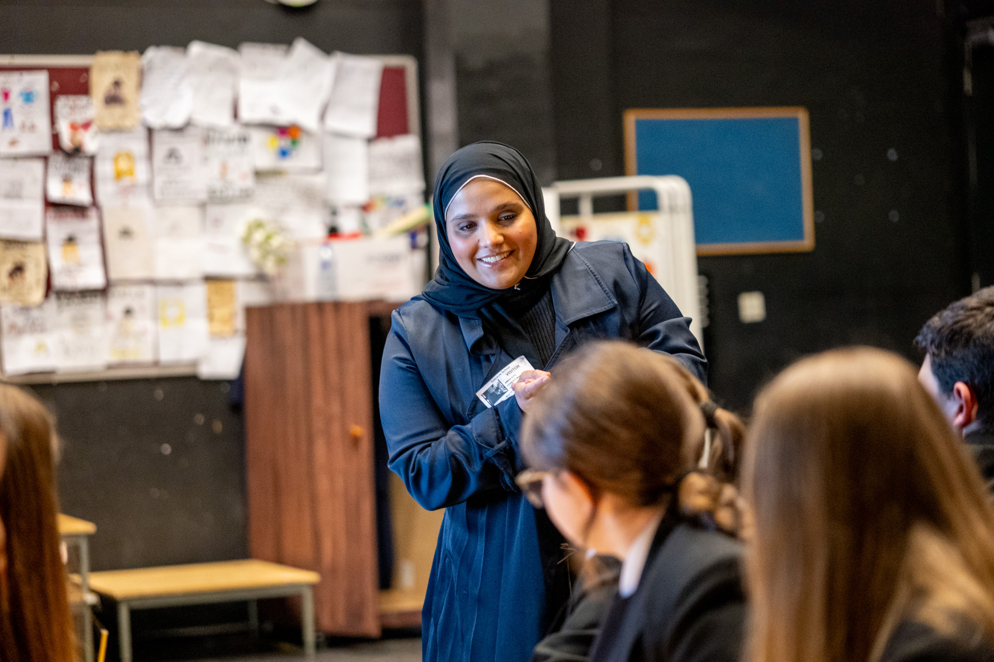 A young woman, stood in front of some children, smiling and looking at them with joy.